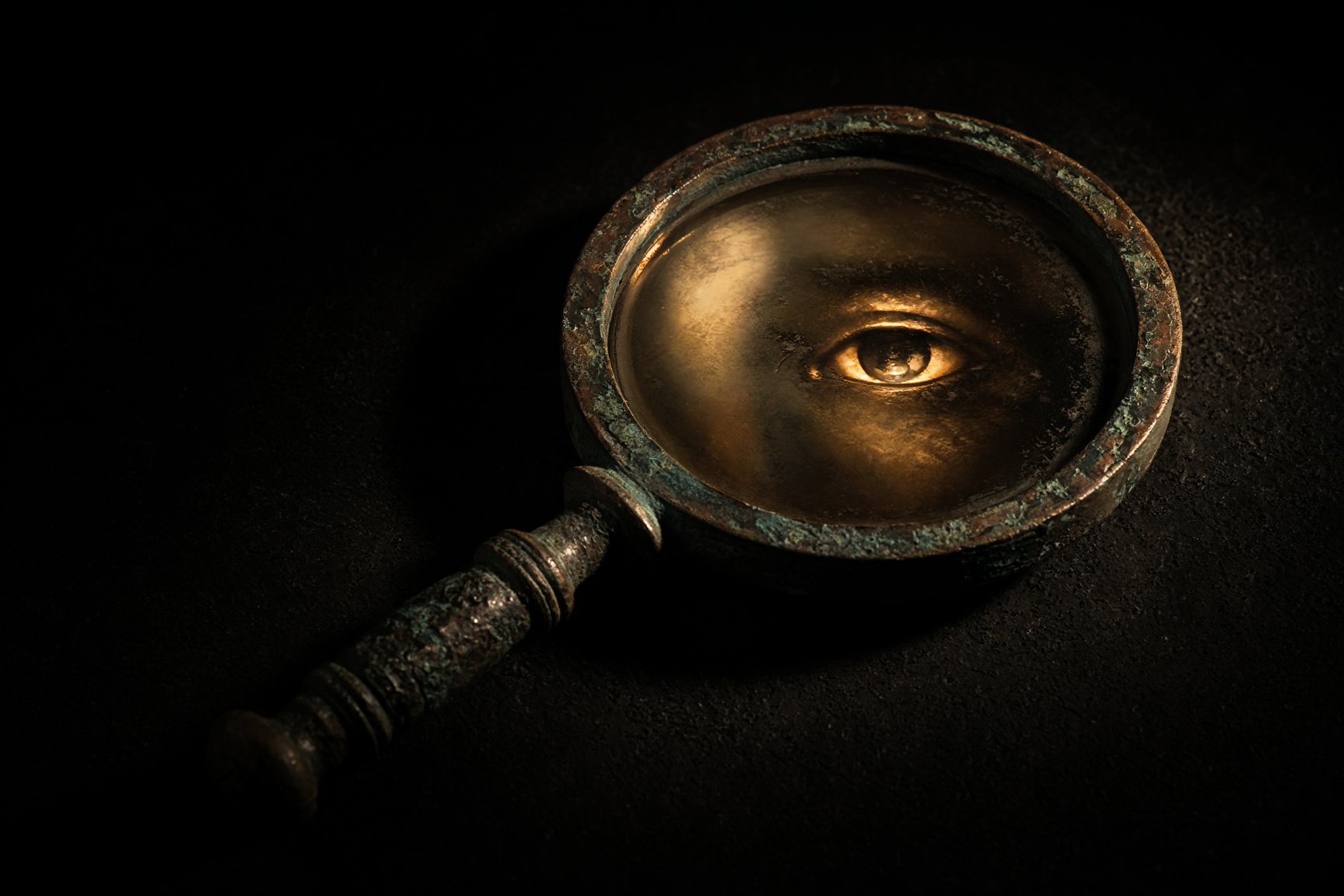 A Roman bronze hand-mirror in darkness, a fragment of an eye in the disc.